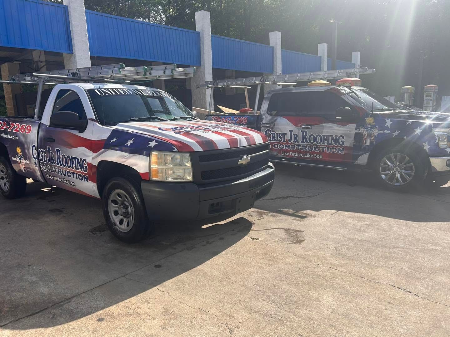 Two patriotic race vehicles parked outdoors in sunlight, one white truck in front and a black car behind.