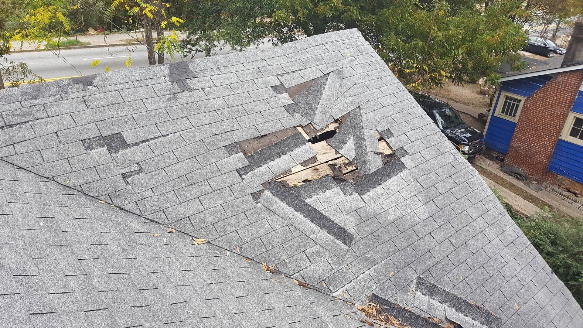 Damaged shingled roof with a large hole and exposed wood near trees and a brick building