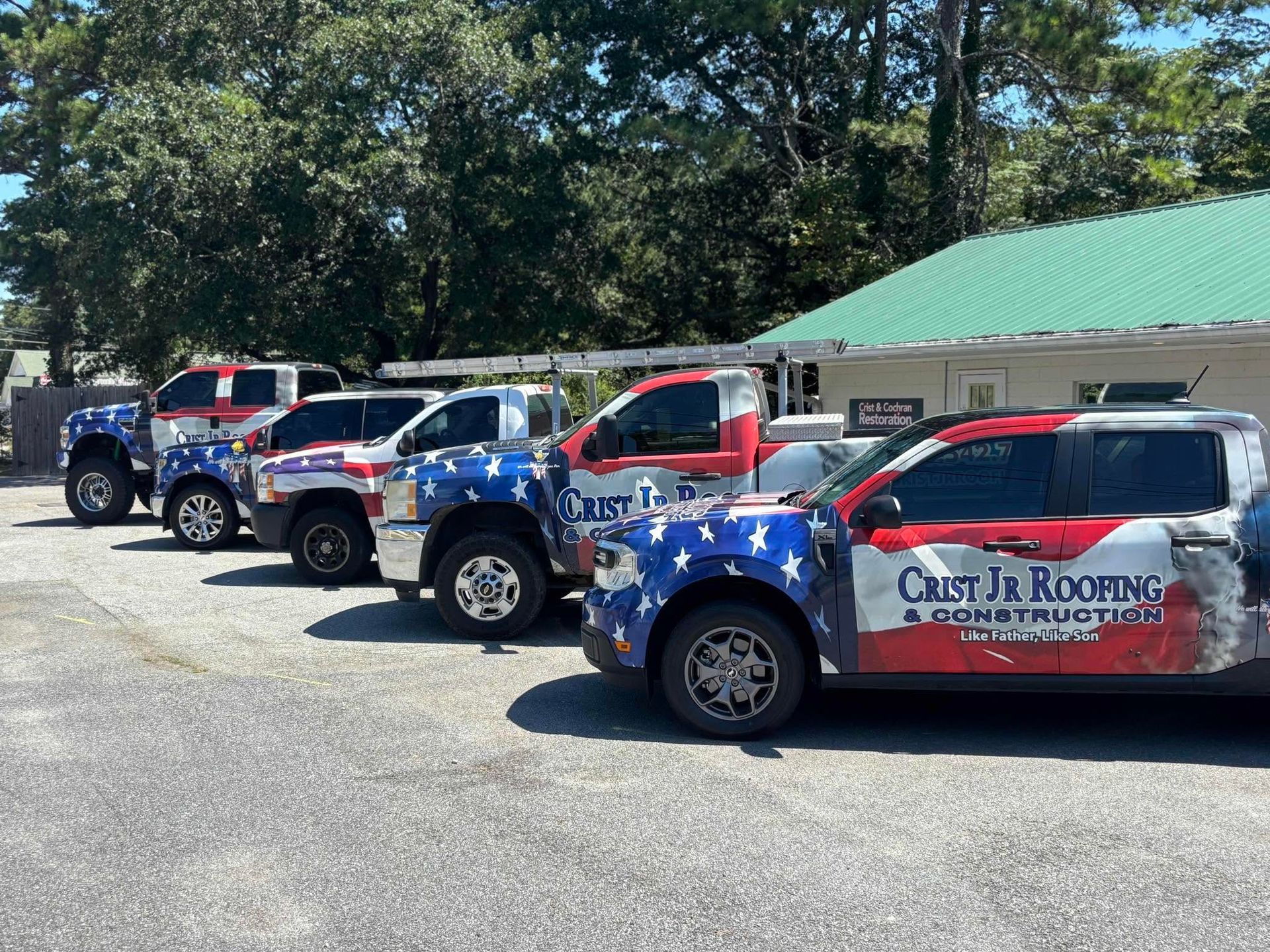 Row of red, white, and blue roofing trucks parked outside a green-roofed building on a sunny day