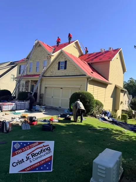 Roofing crew working on a tan house with red shingles, tools and materials on the lawn