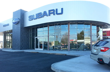 A modern Subaru car dealership with large glass windows and white signage under a bright sky, with a car in the foreground.