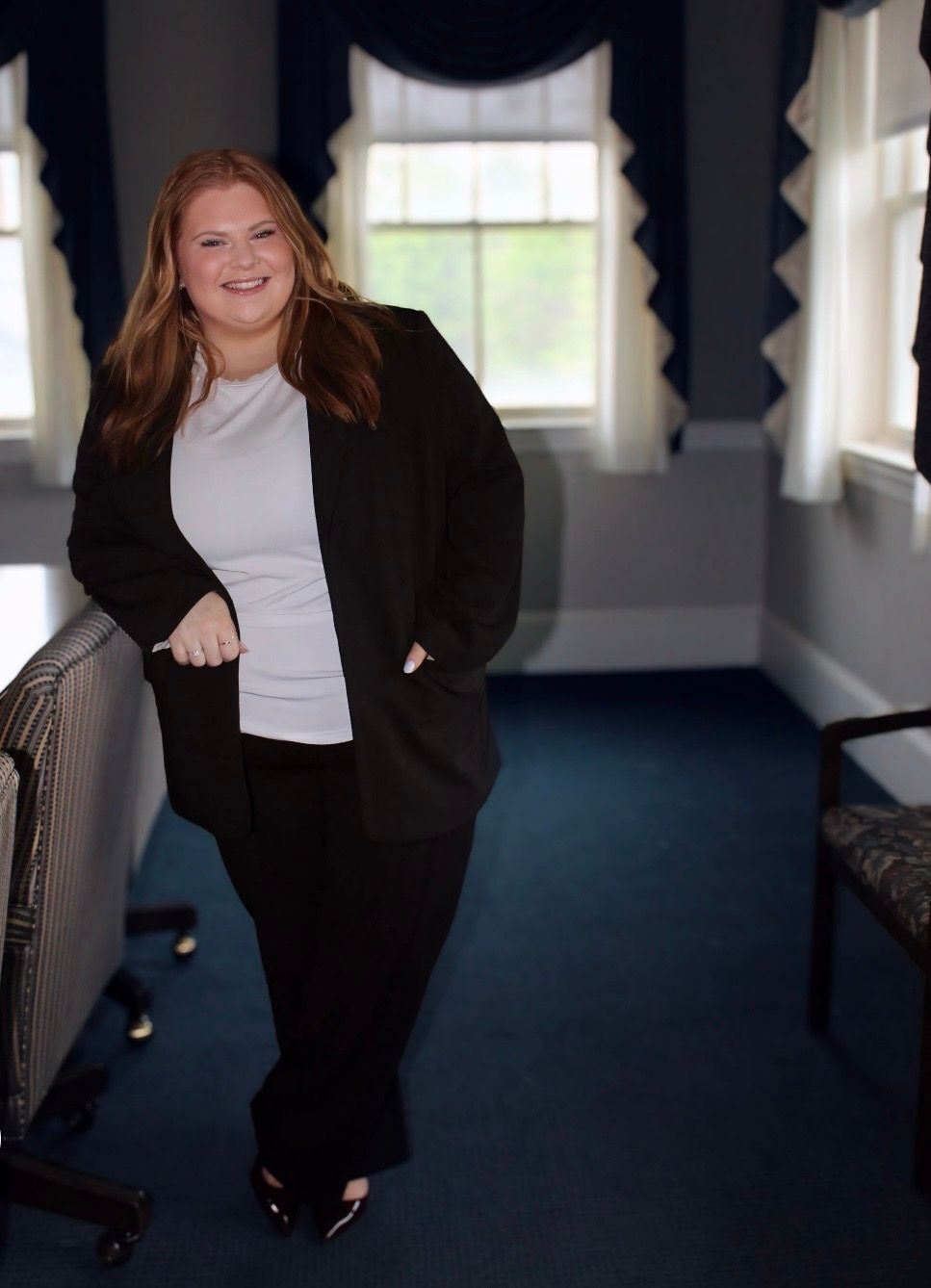 Woman in a black suit leaning in a hallway with dark blue walls and bright windows.