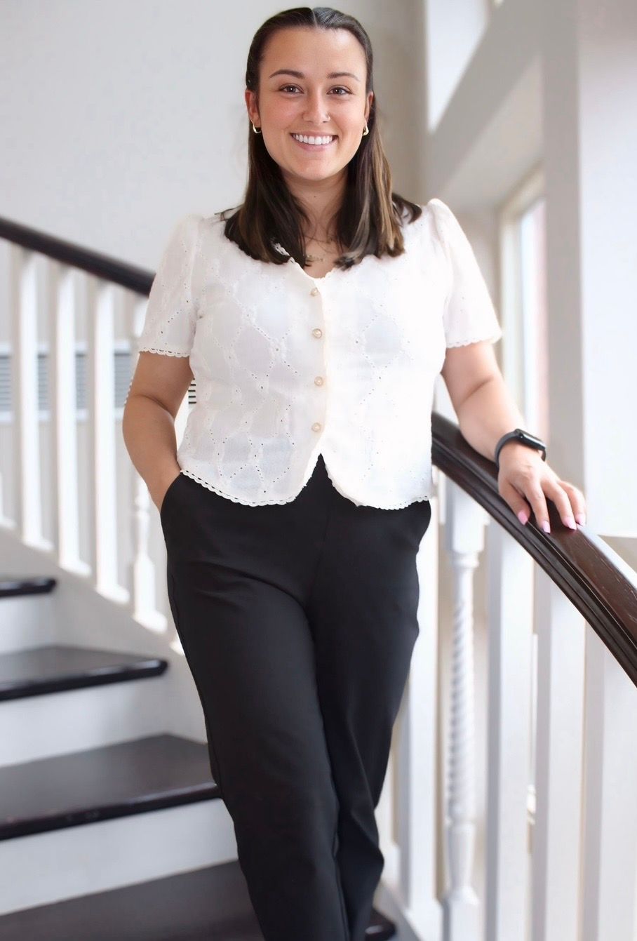 Woman standing on a staircase, smiling, wearing a white blouse and black pants.