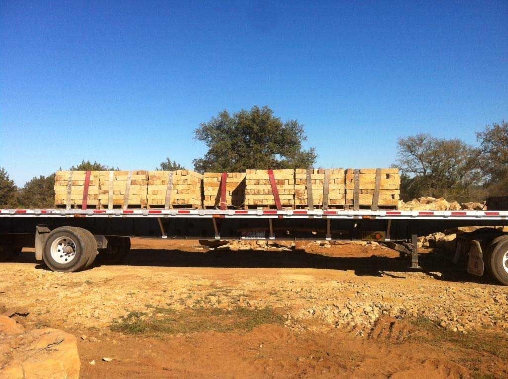 Flatbed truck carrying strapped stacks of tan-colored blocks on a dirt road, under a blue sky.