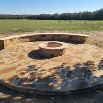 Circular stone patio with fire pit and curved seating, set in a grassy field.
