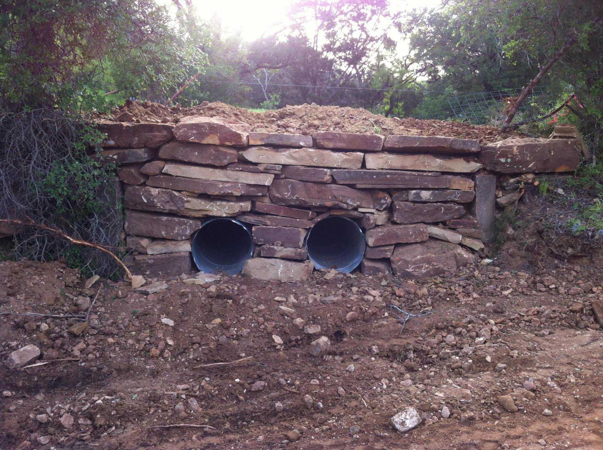 Stone-lined culvert with two metal pipes. Dirt ground. Dense green foliage in the background.