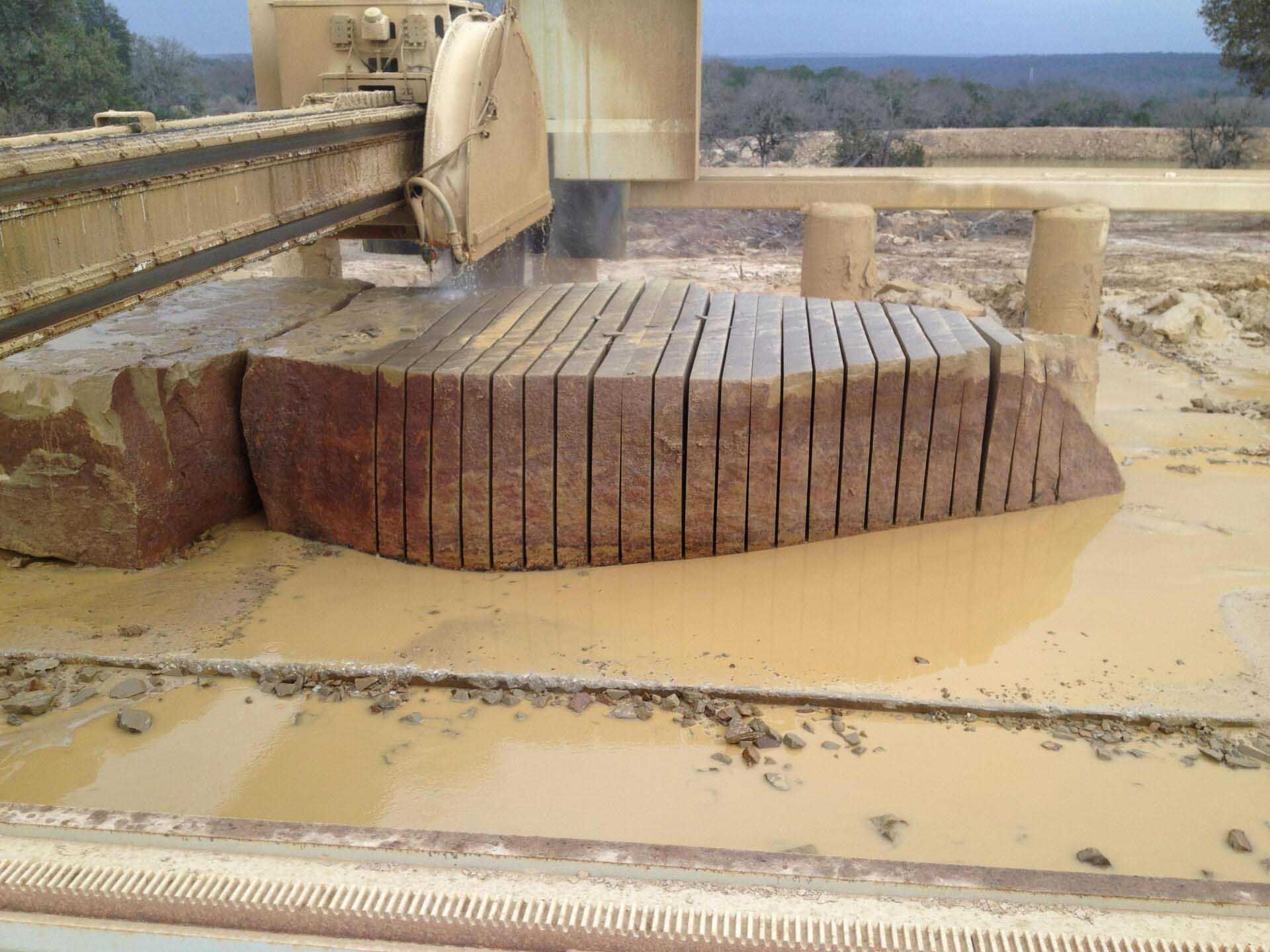 A large stone being cut into slabs by a machine in an outdoor setting. Brown stone and beige machinery.