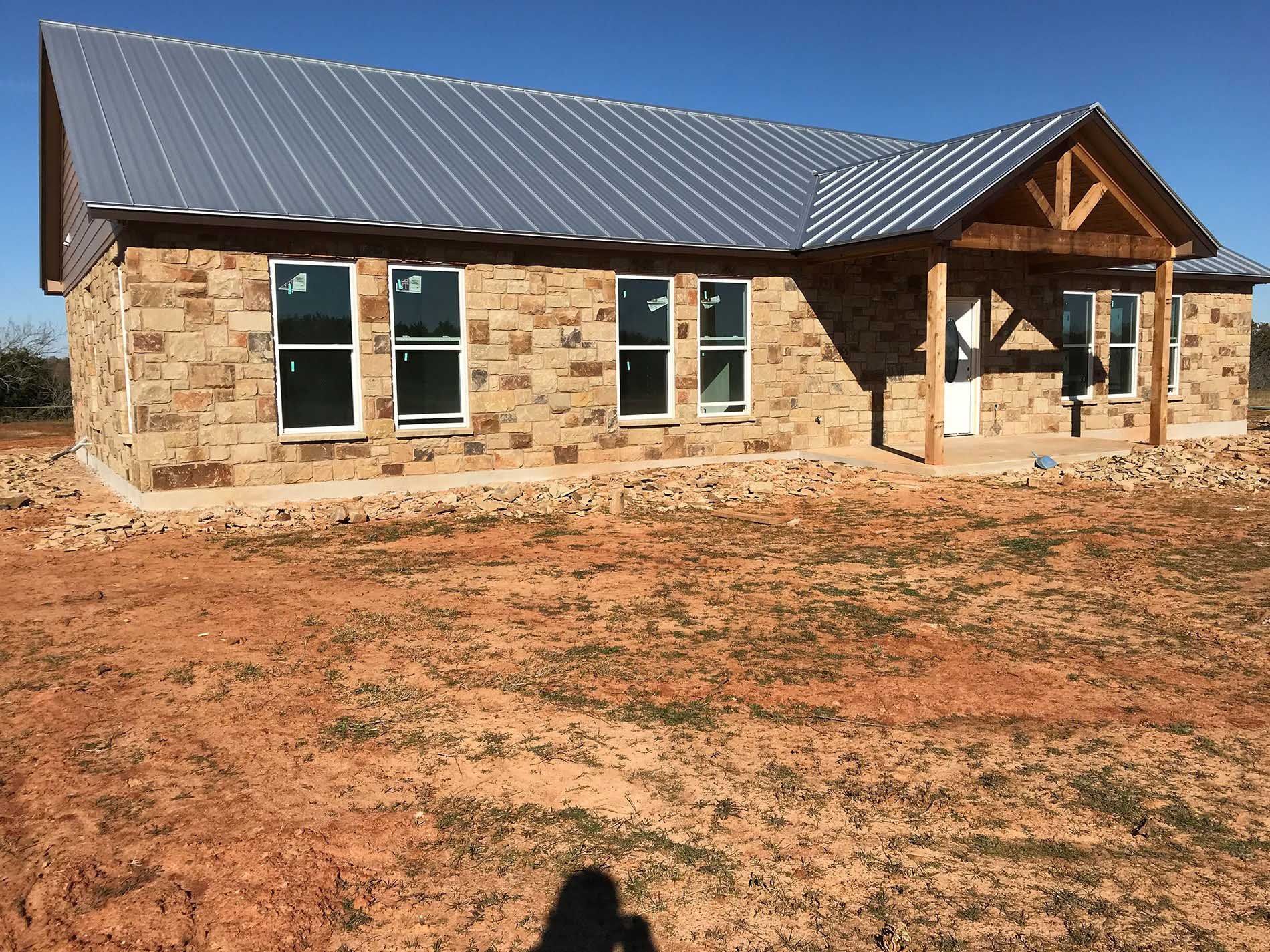 Tan brick home with metal roof, small porch, and open yard under blue sky.