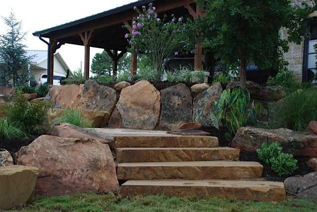 Stone steps lead up to a covered porch; large boulders surround the stairs, with landscaping.