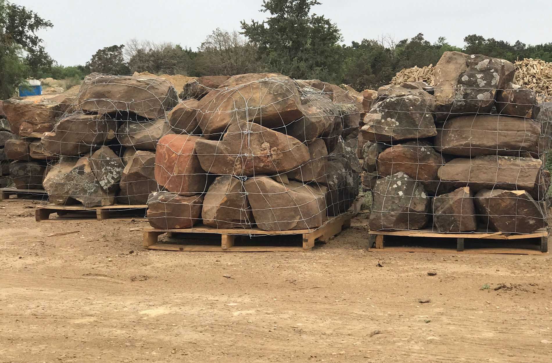 Three pallets of large rocks, bundled with wire, on dirt ground.