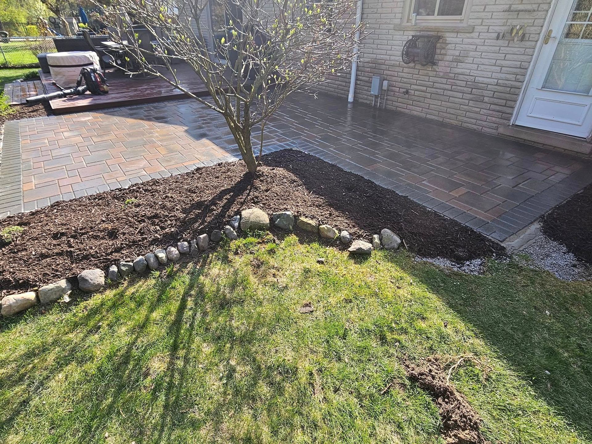 A backyard patio with a small tree in the center, surrounded by mulch and a rock border.