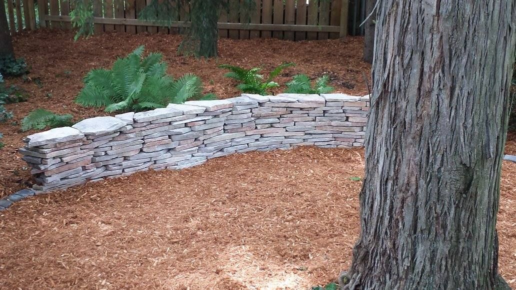 Stone retaining wall curves through a wood-chipped garden, backed by a wooden fence. A large tree trunk is in the foreground.