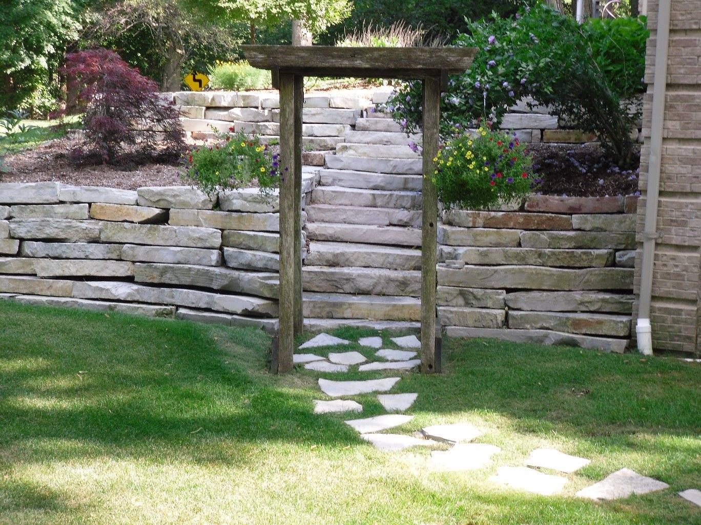 Wooden archway frames stone steps ascending a terraced garden, with a flagstone path leading to it from a grassy lawn.