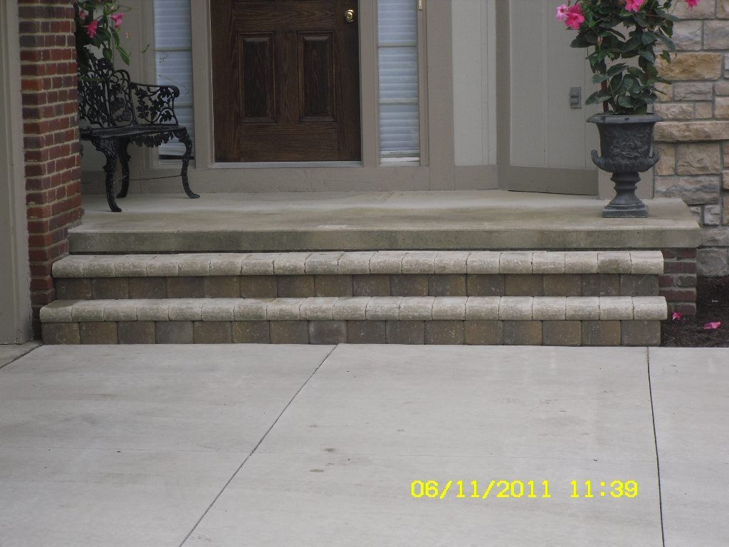 Three-step concrete entrance with brick accents, leading to a wooden door; flower pots flank the entrance.