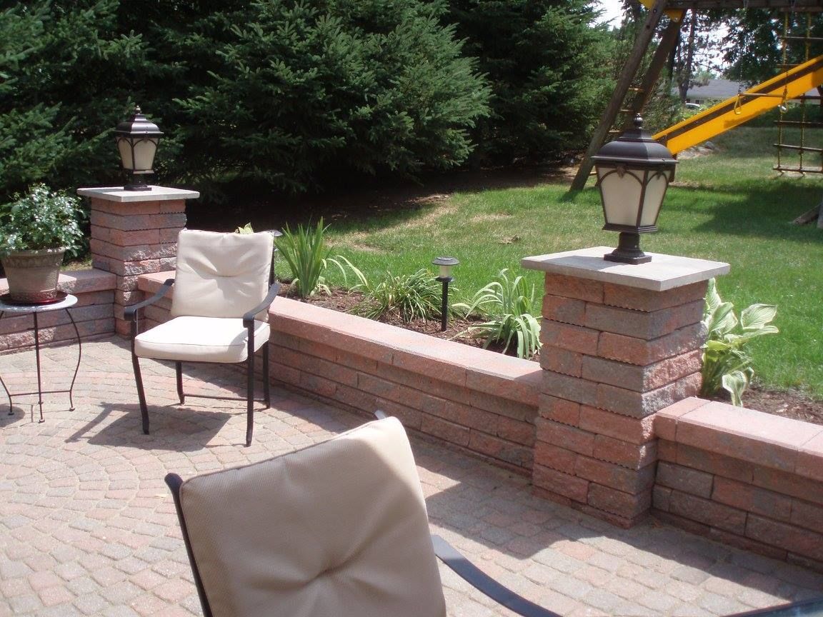 A brick patio with two chairs, a low wall, and two pillars with lamps. Green grass and trees are in the background.