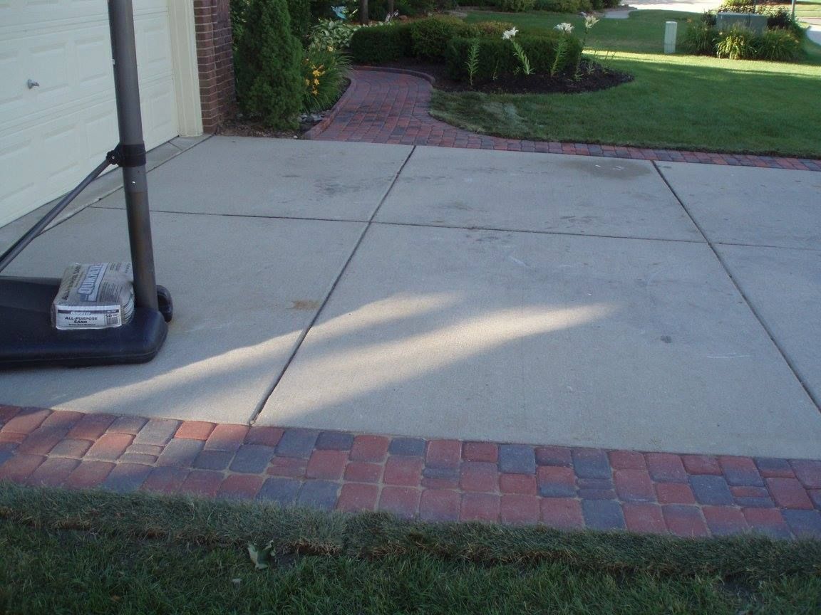 Driveway with red brick border, basketball hoop base, and adjacent green lawn and flower bed.