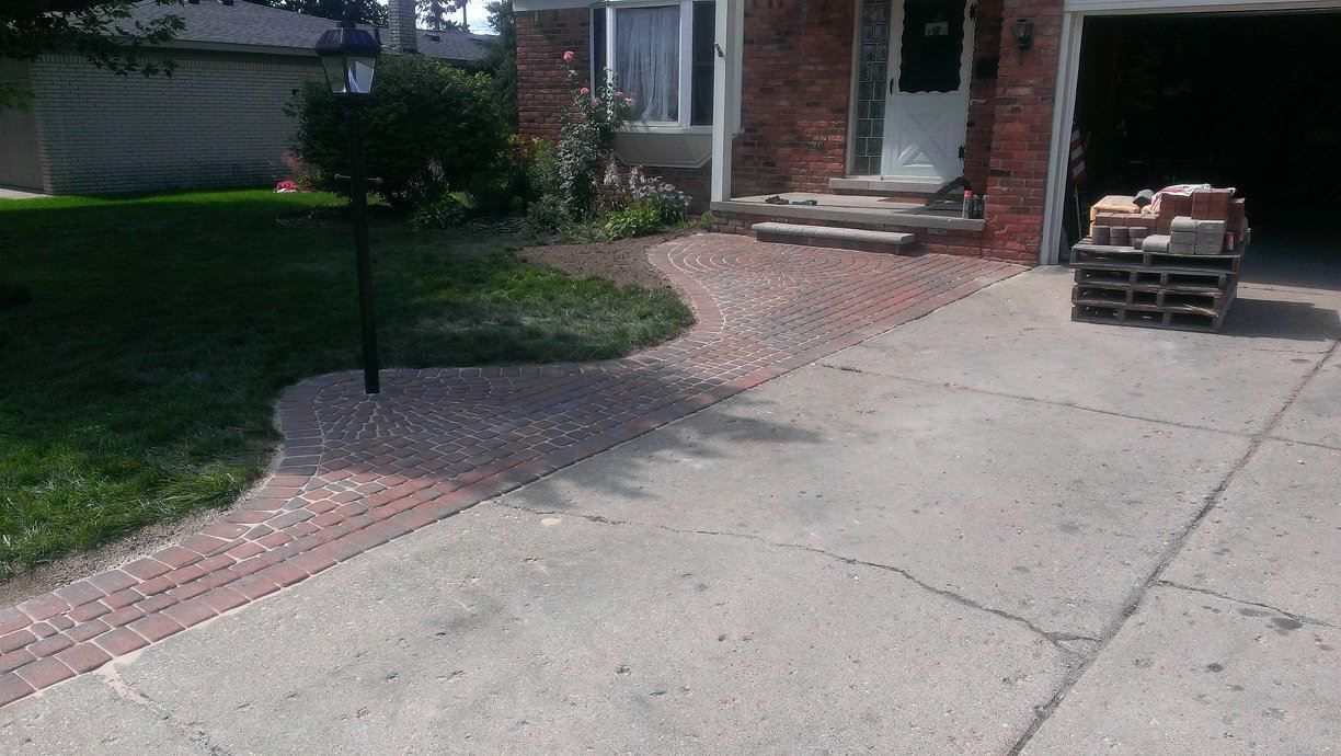 Concrete driveway with a red brick walkway leading to the front steps of a brick house.  Pallet of bricks sits near the garage.