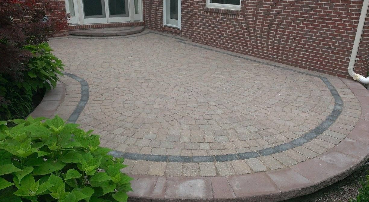 Gray house with a paved driveway, stone pillars, and a clear blue sky. Green trees and lawn surround the property.