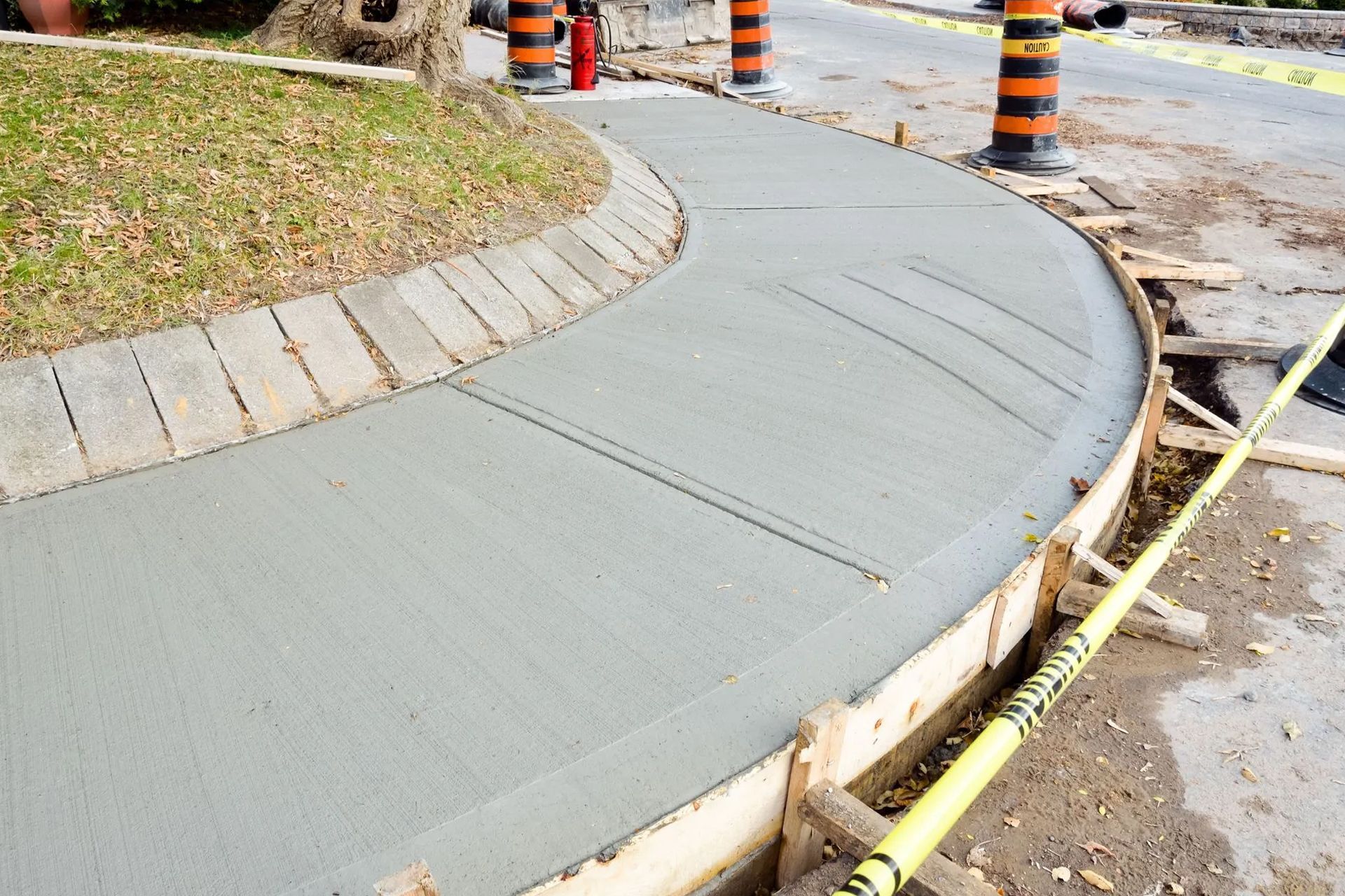 Freshly poured concrete sidewalk under construction, curved around a grassy area, with wooden forms and safety cones.