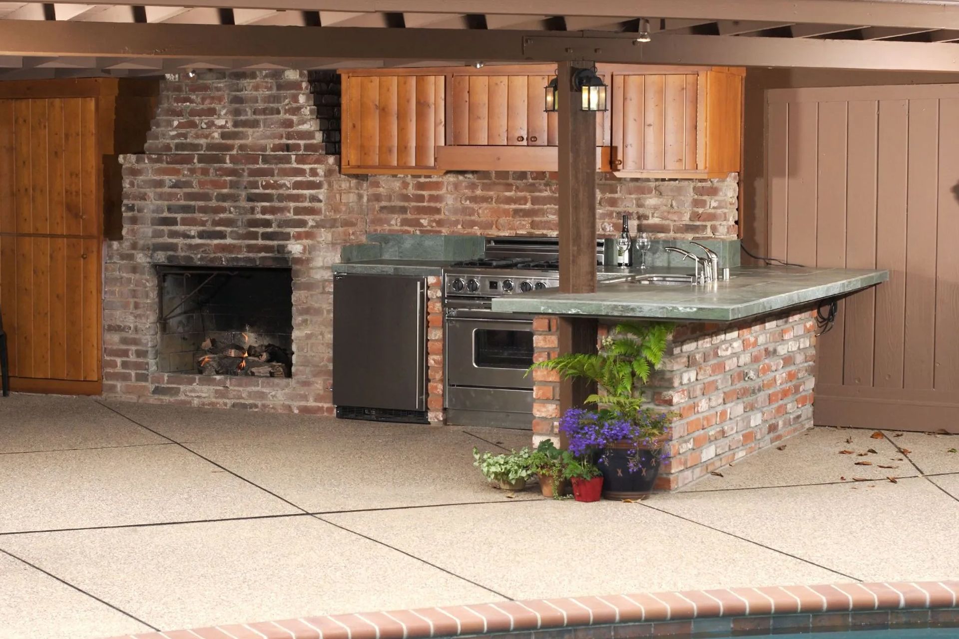 Outdoor kitchen with brick fireplace, cooking area, and countertop with a pool in the foreground.