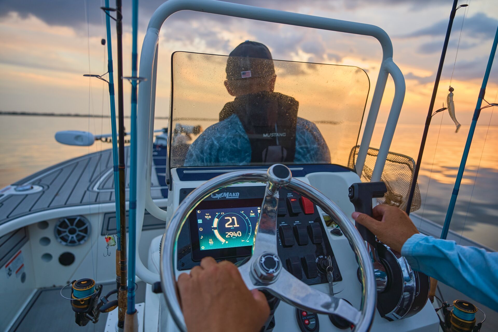 A person steers a fishing boat at sunset, with a digital display showing 21 mph and fishing rods visible on the deck.