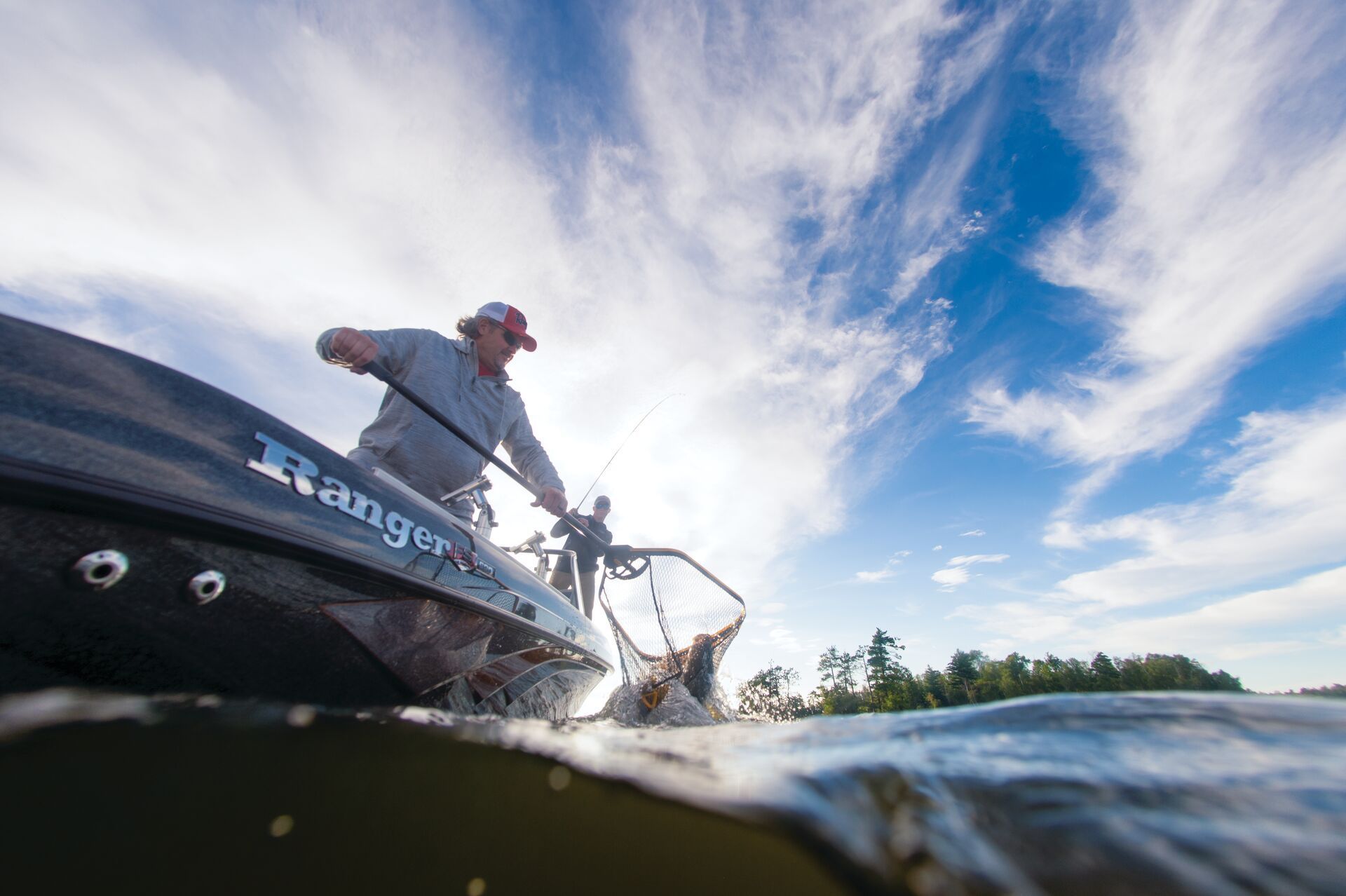 A silver and black fishing boat on a trailer parked on gravel, with green trees in the background.