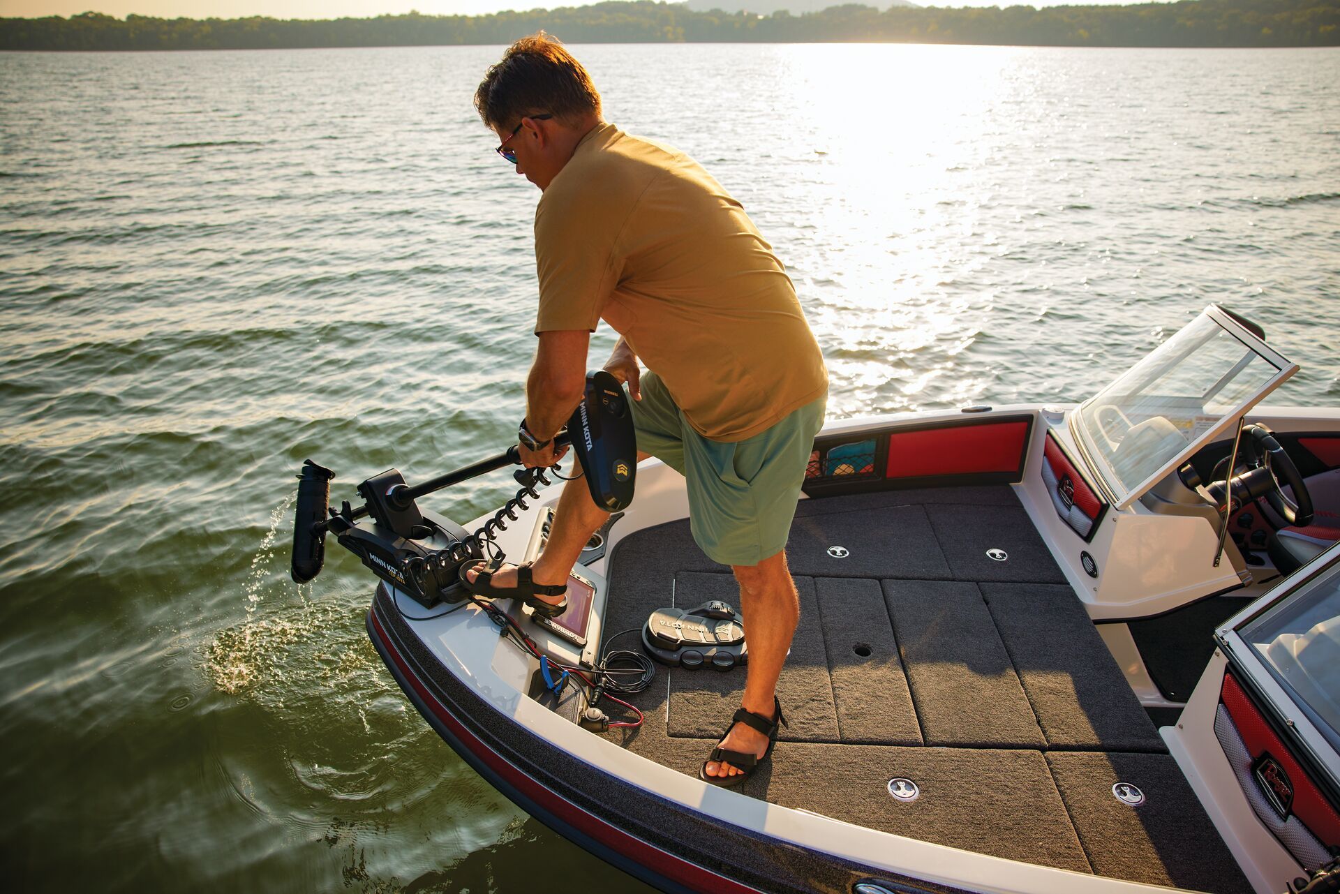 A person standing on the bow of a boat at sunset, preparing to deploy a mounted trolling motor into the water.