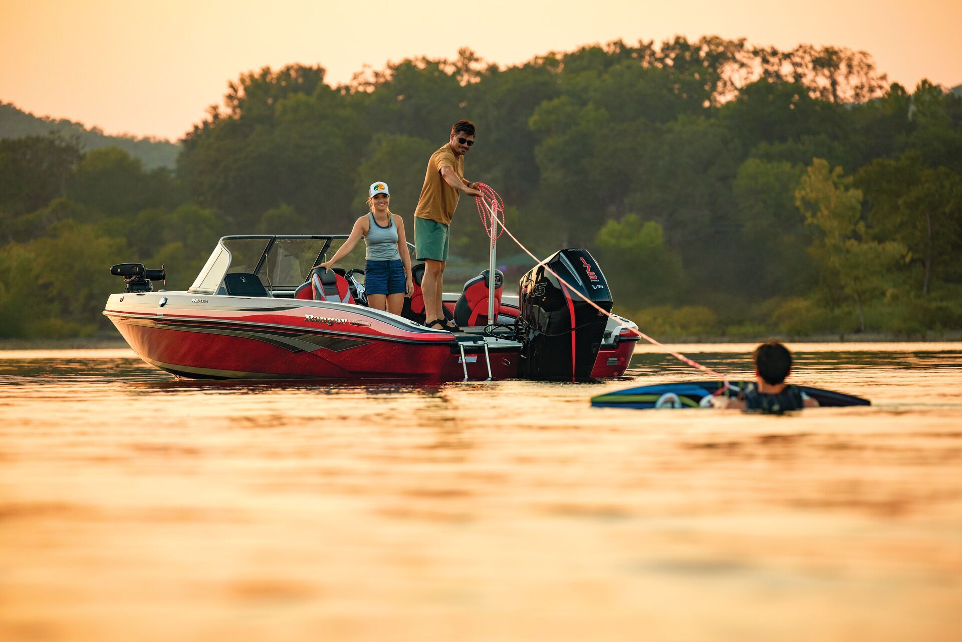 A red motorboat with two people on board prepares to pull a swimmer on a wakeboard on a calm lake at sunset.