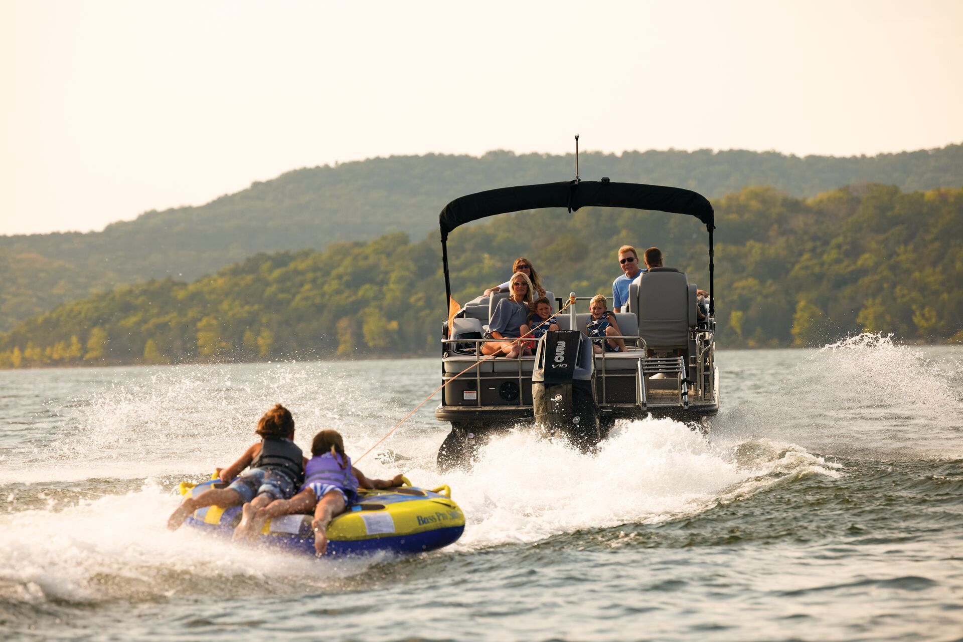 Two people ride an inflatable tube being pulled by a pontoon boat on a sunny lake with hills in the background.