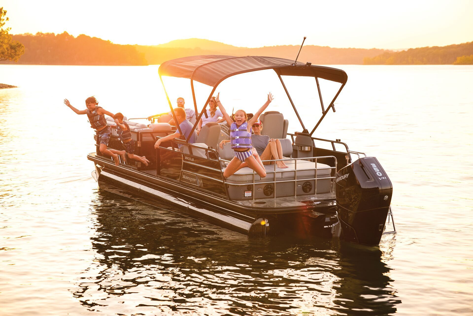 People jump from a black pontoon boat into a lake at sunset.