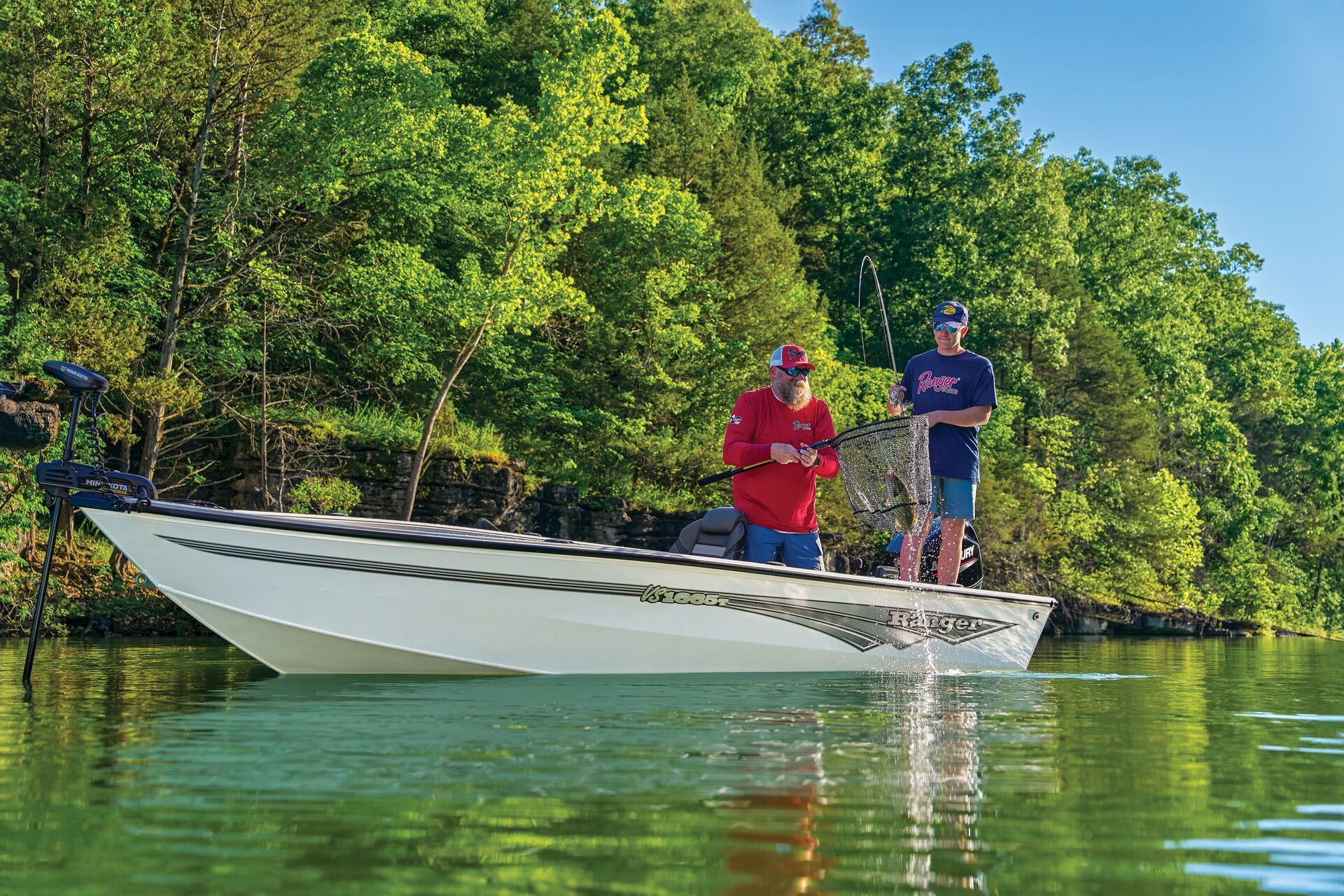 Two people stand on a white boat in a calm lake, surrounded by lush green trees on a sunny day.