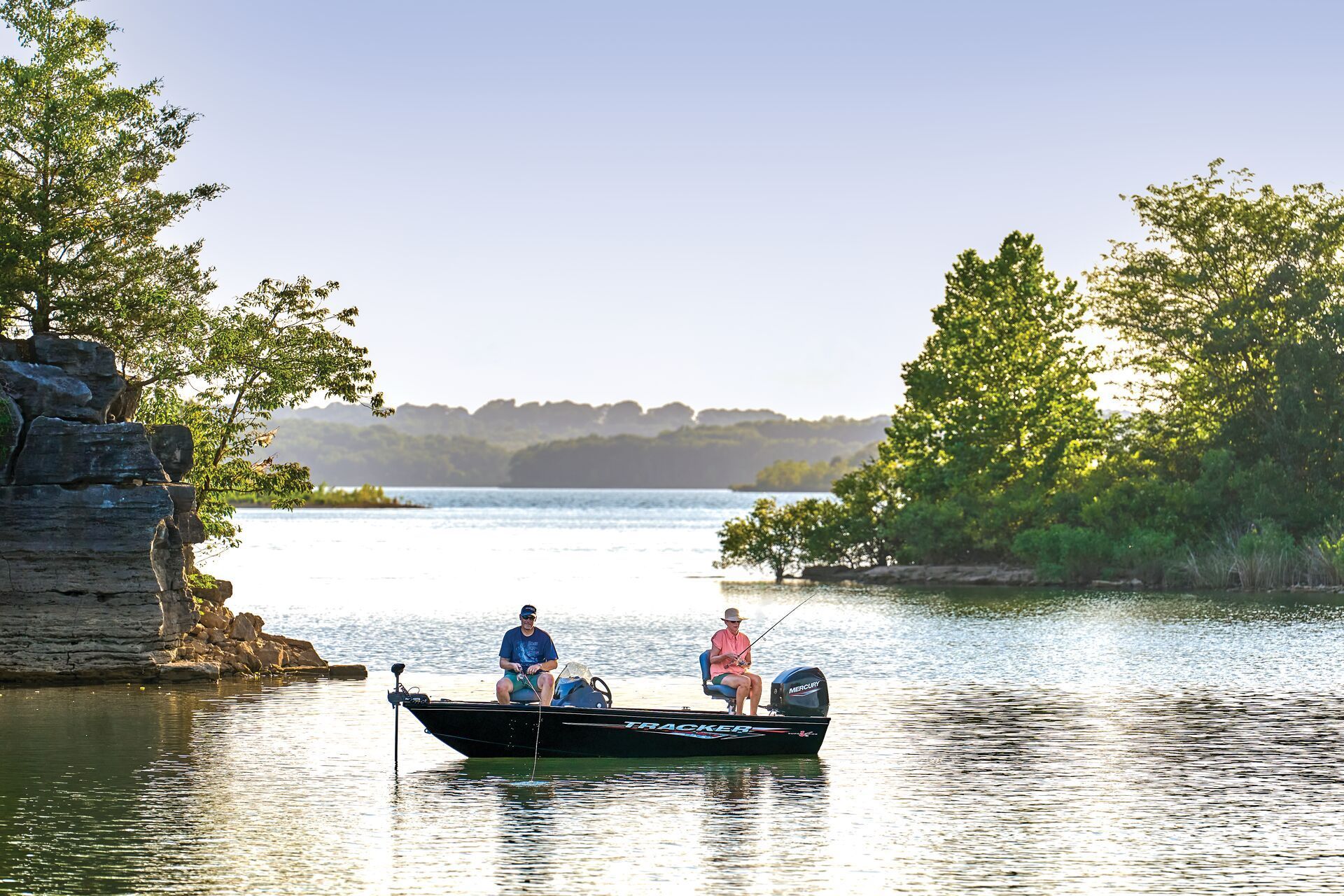 Two people sit in a small black motorboat fishing on a calm lake surrounded by trees and rock formations.