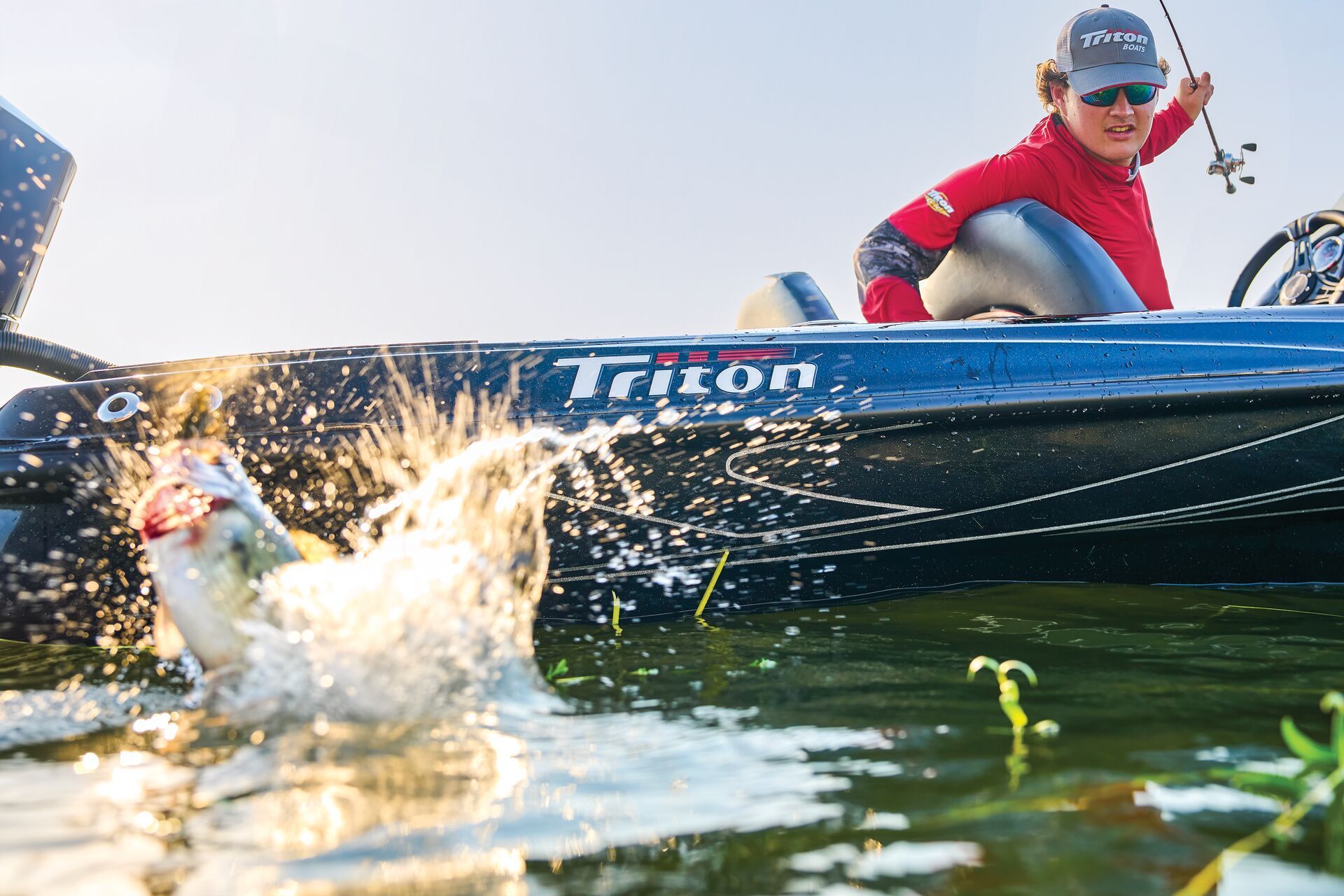 A person wearing a red shirt and cap on a blue Triton boat reels in a fish splashing near the water's surface.