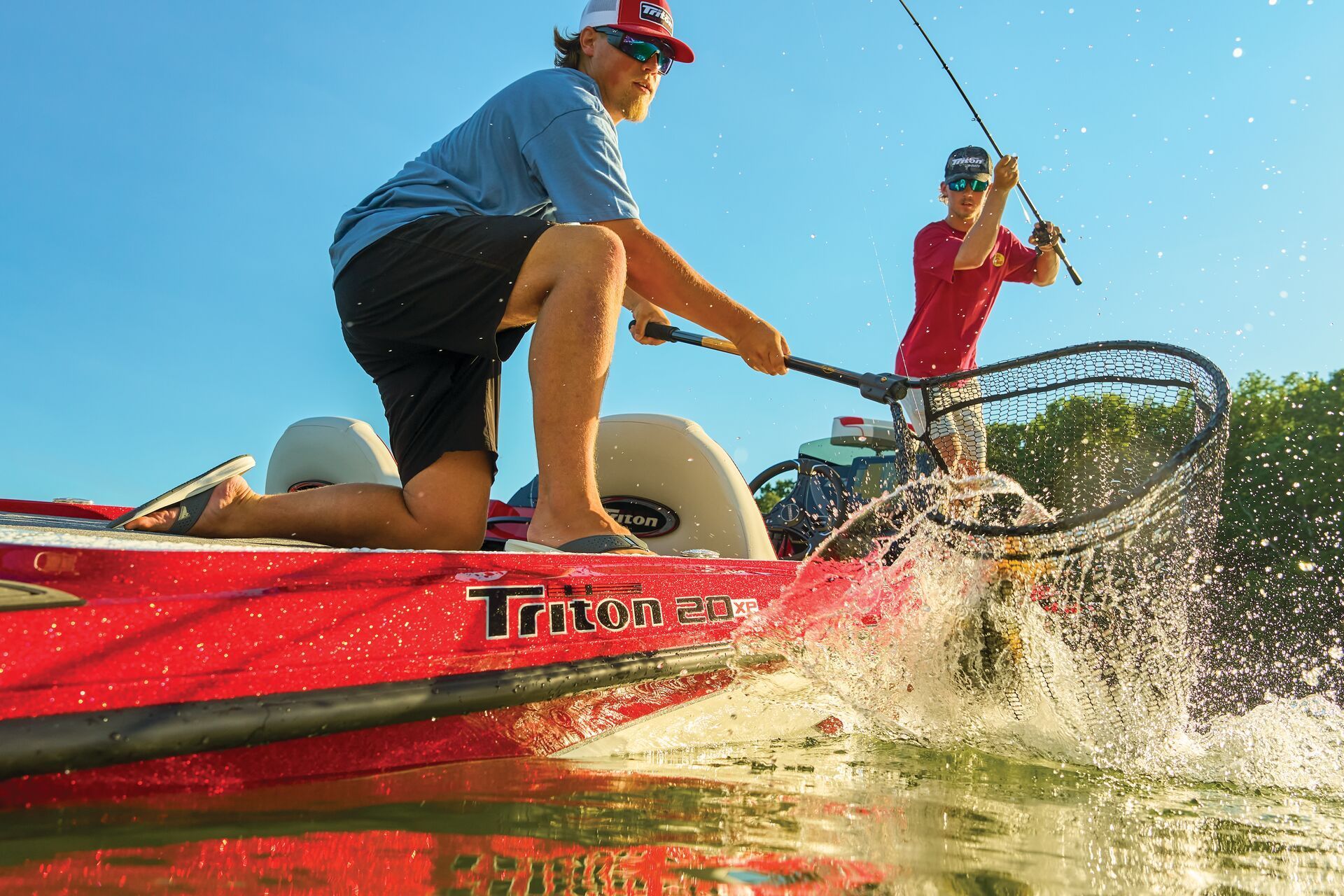 Two people in a boat on sunny water, one using a landing net to catch a fish while the other holds a fishing rod.