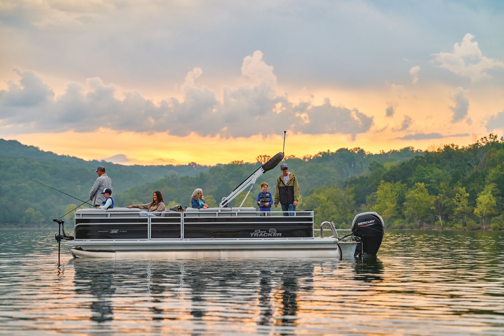 A group of people fishing from a pontoon boat on a calm lake during a golden sunset, with forested hills in the background.