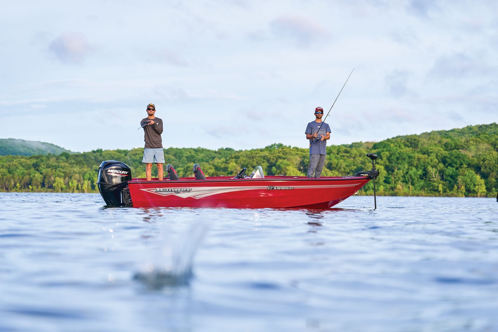 Two people stand on a bright red boat on a calm lake, surrounded by green hills, with a water splash in the foreground.