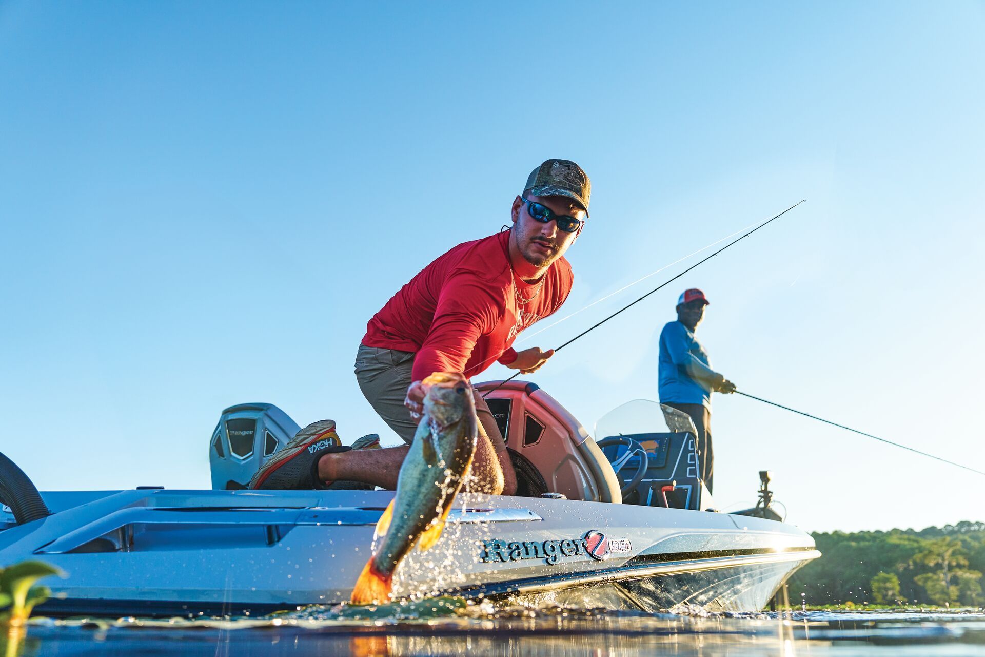 A person in a red shirt kneels on a boat, holding a fish, while another person fishes in the background under a blue sky.