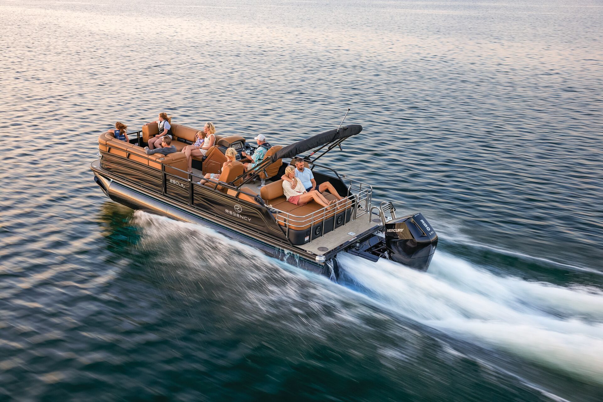 A group of people sitting on a moving brown and black pontoon boat on a blue lake, with a white wake trailing behind.