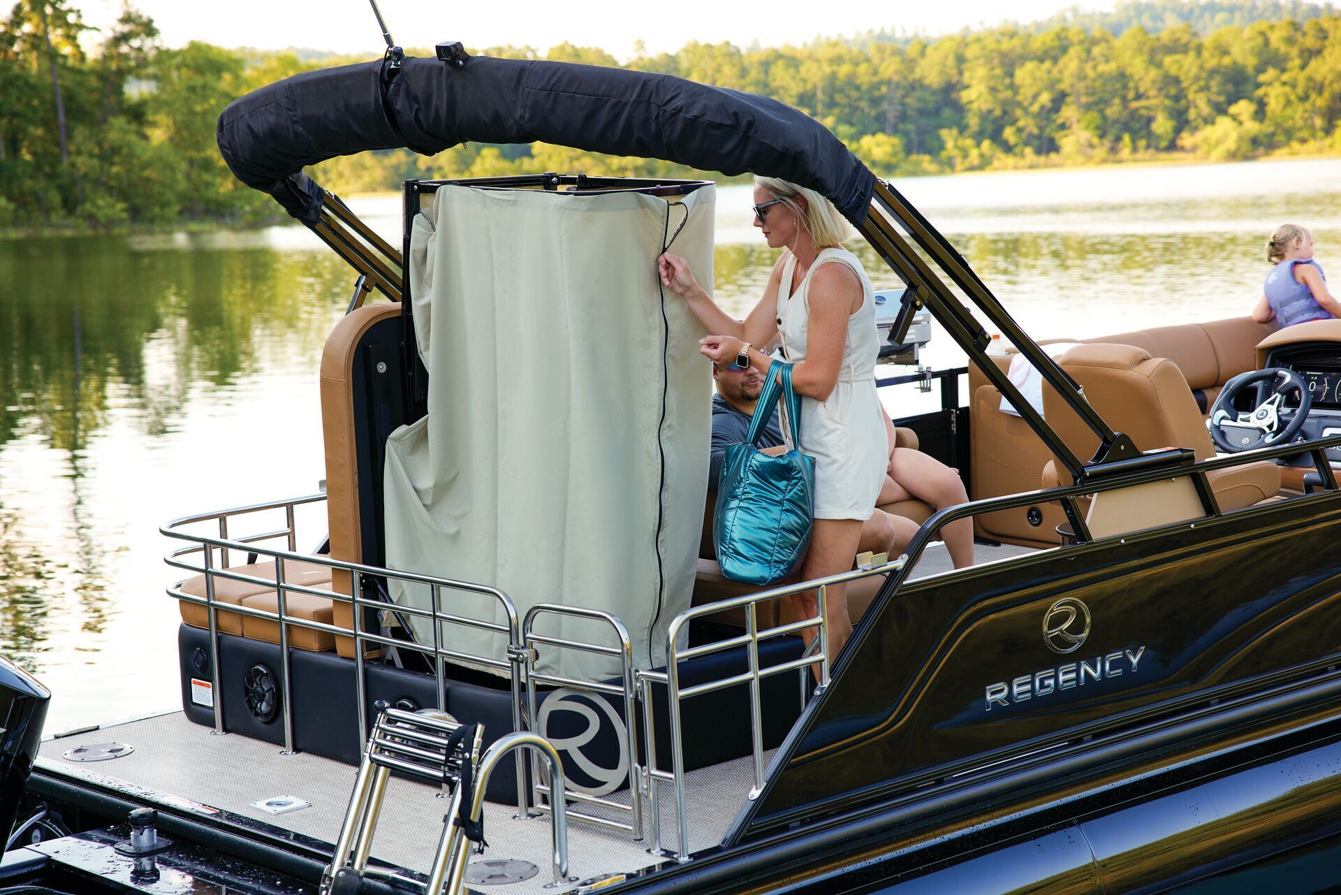 A person stands on a black pontoon boat, adjusting a beige privacy curtain enclosure under the canopy.