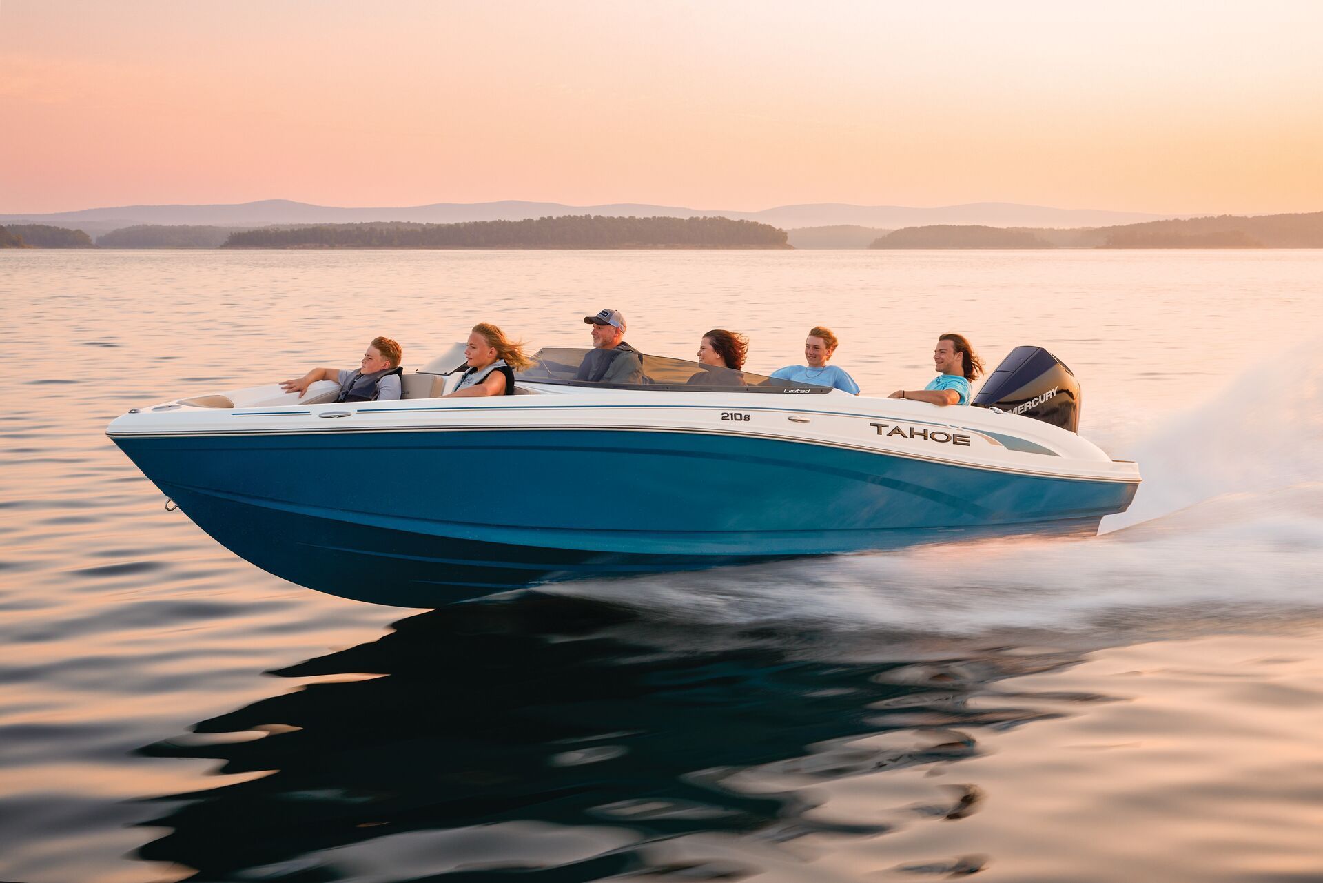 A blue and white motorboat speeds across a calm lake at sunset with six people sitting on board.