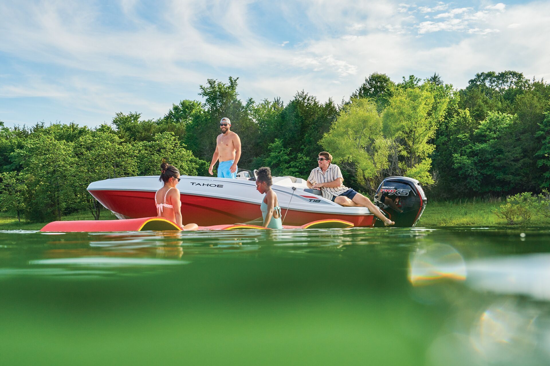 Four people relax on a floating mat next to a red and white motorboat on a sunny lake surrounded by trees.