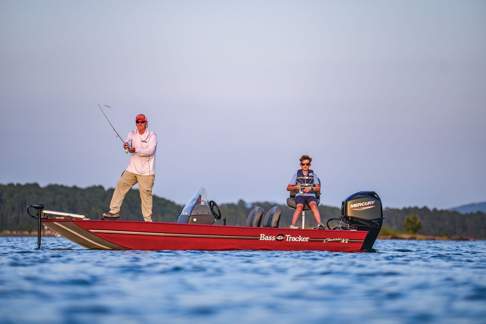 An adult and a child in life jackets fishing from a red motorized boat on calm water with a forest in the background.
