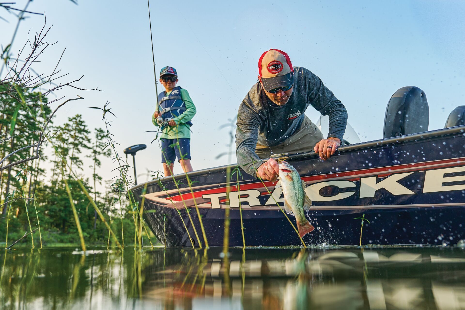 A person in a fishing boat is lifting a caught fish out of the water while a second person fishes in the background.