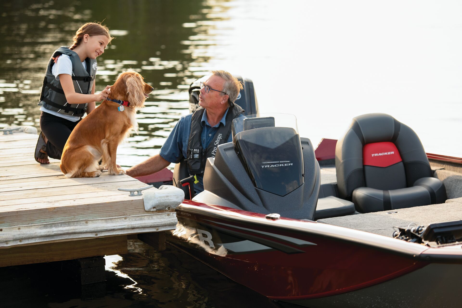 A person and a child wearing life jackets interact with a golden dog on a dock next to a parked motorboat.