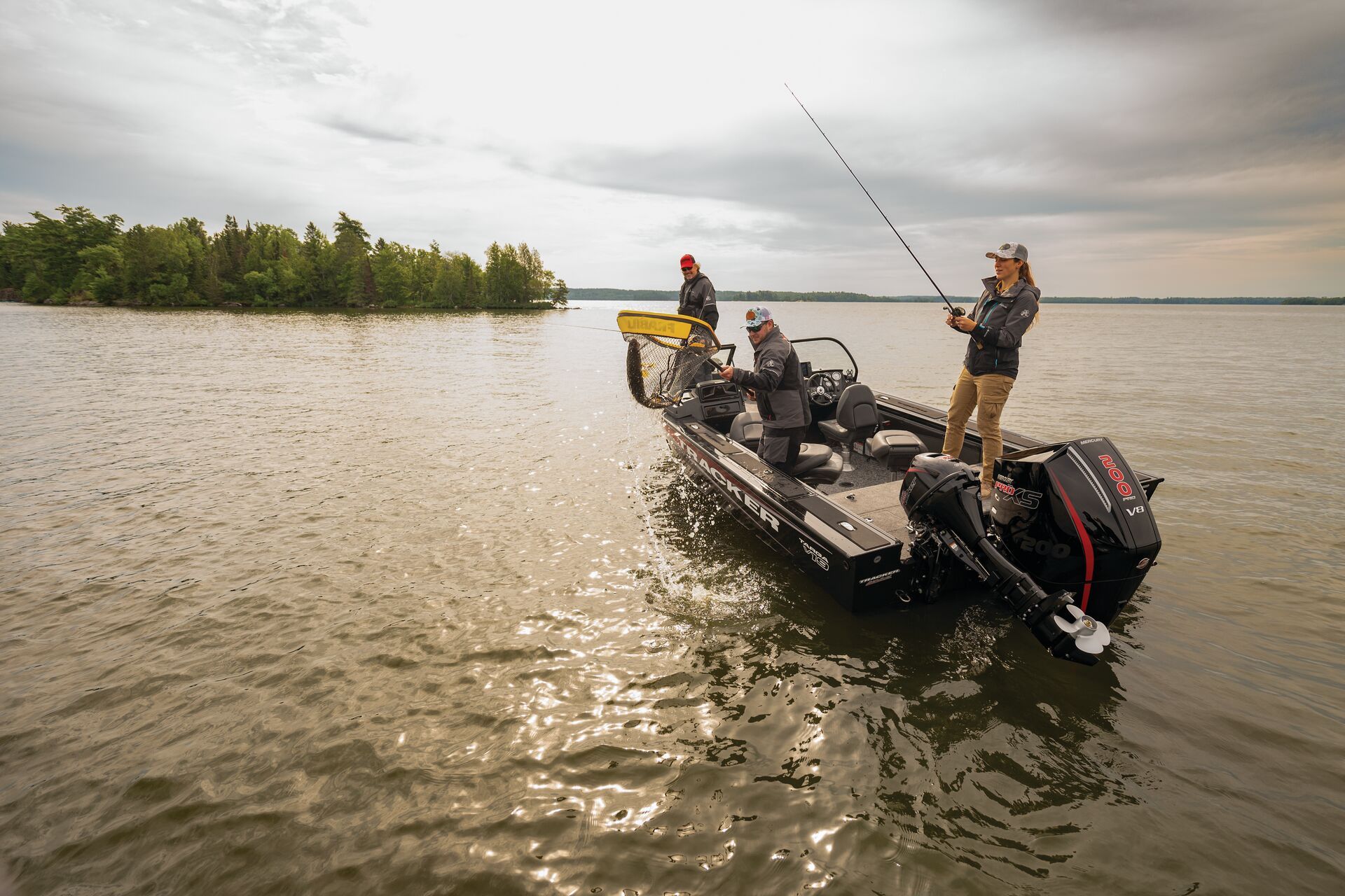 Three people fishing from a motorboat on a cloudy day near a forested shoreline.
