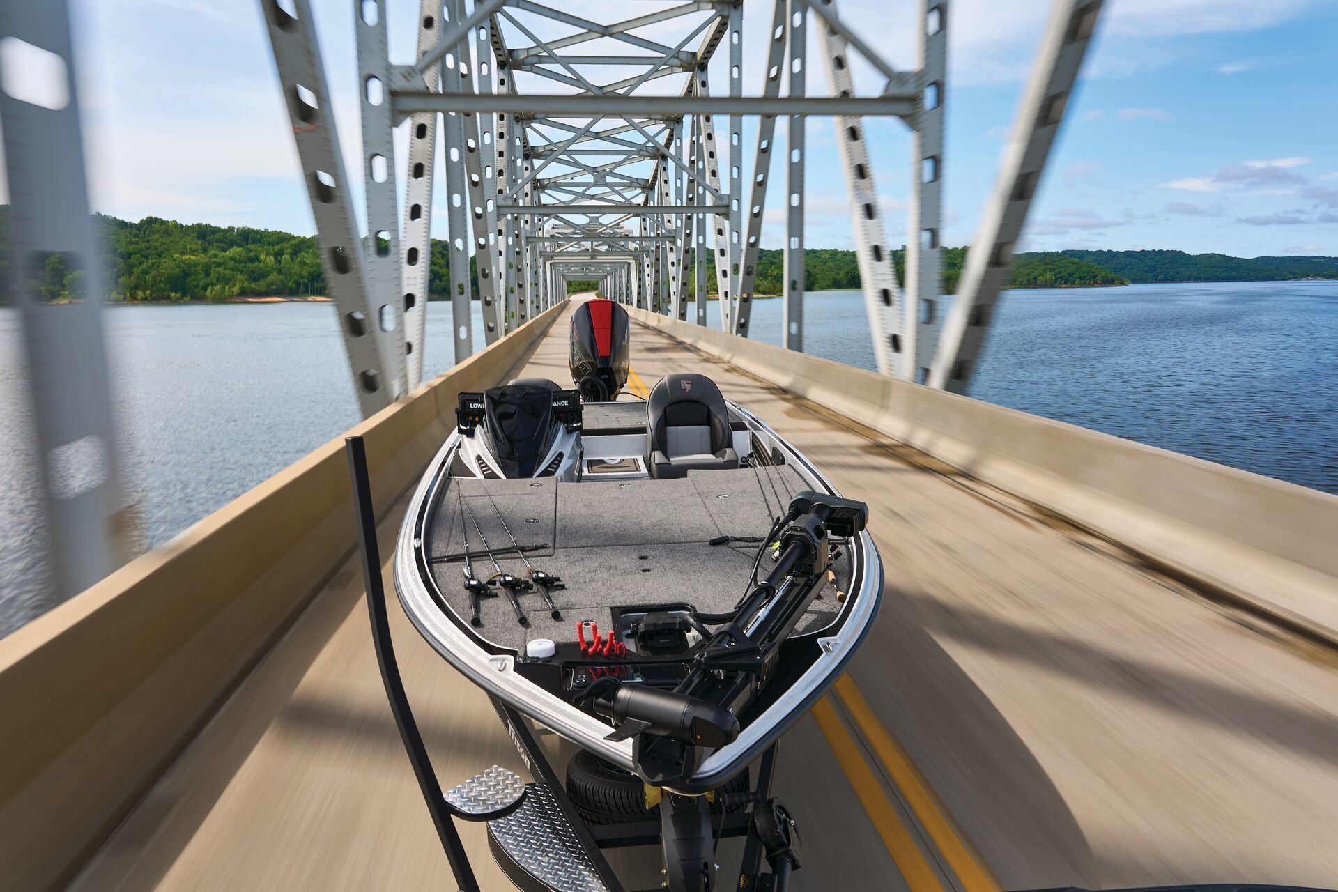 A bass boat being towed on a trailer across a metal truss bridge over a wide, blue lake under a clear sky.