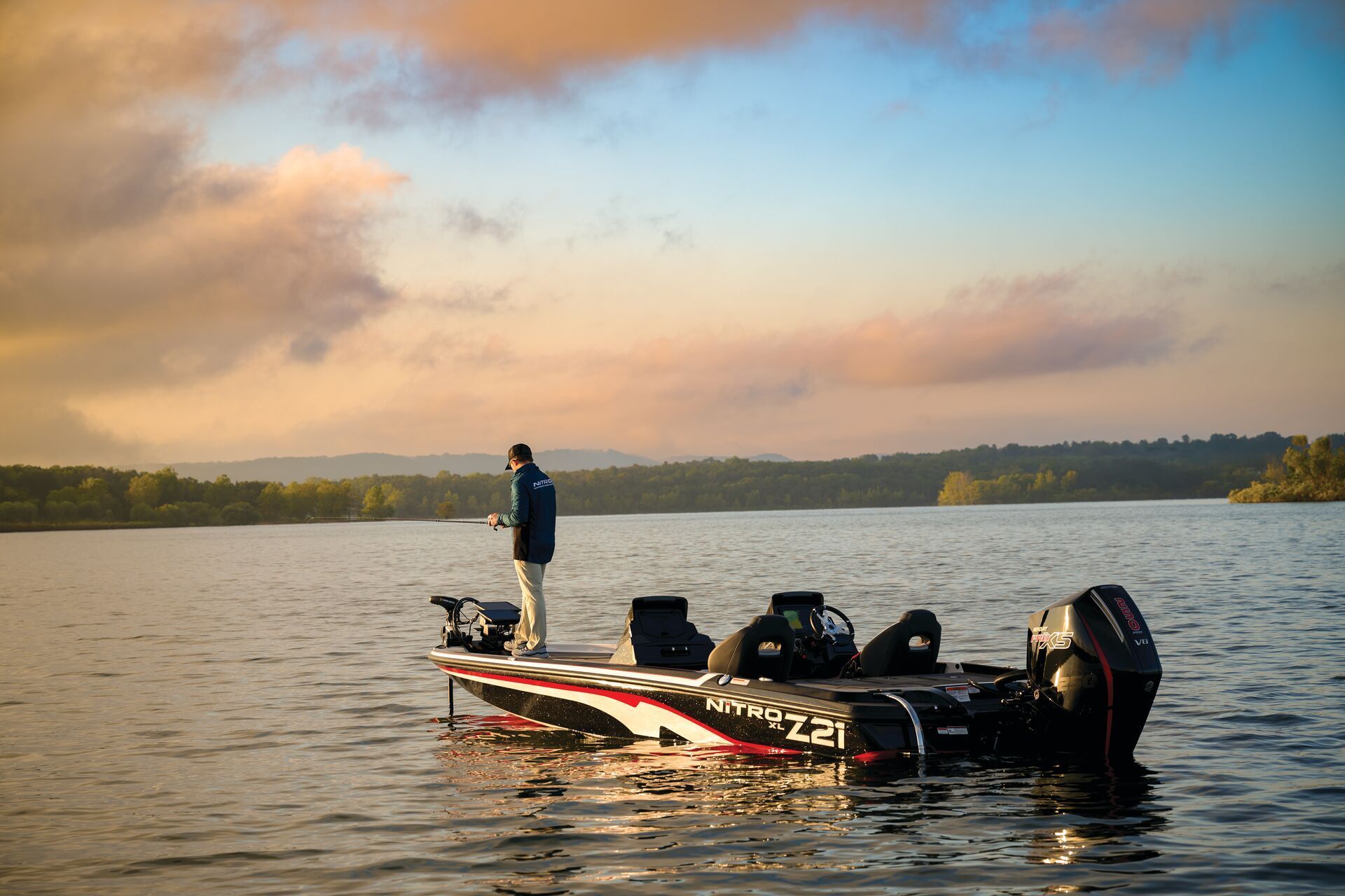 A person fishes from a bass boat on a calm lake at sunrise with a distant treeline under a soft, cloudy sky.