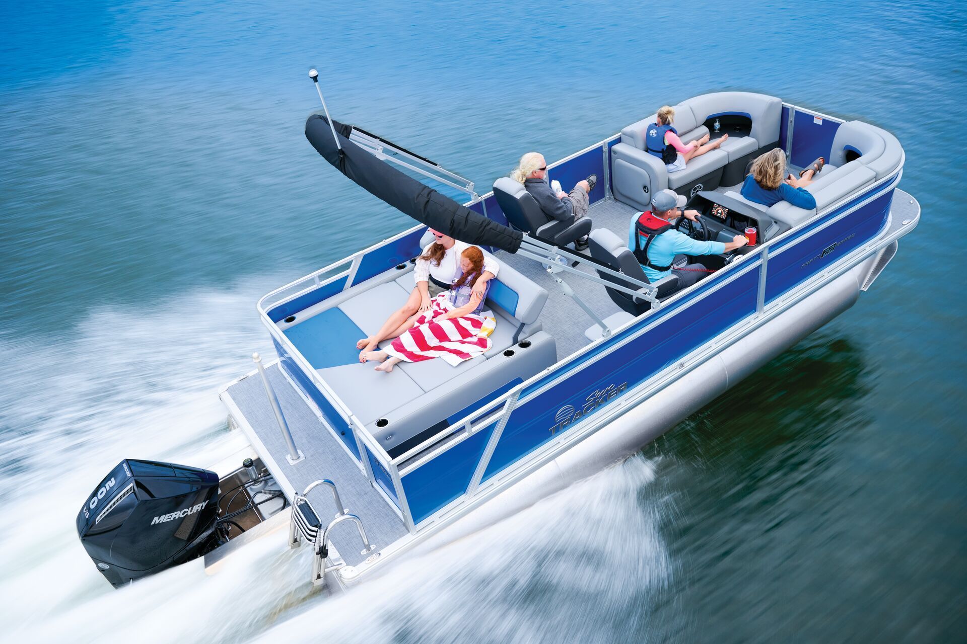 A blue and gray pontoon boat travels across open water with several people on board, featuring a Mercury outboard motor.