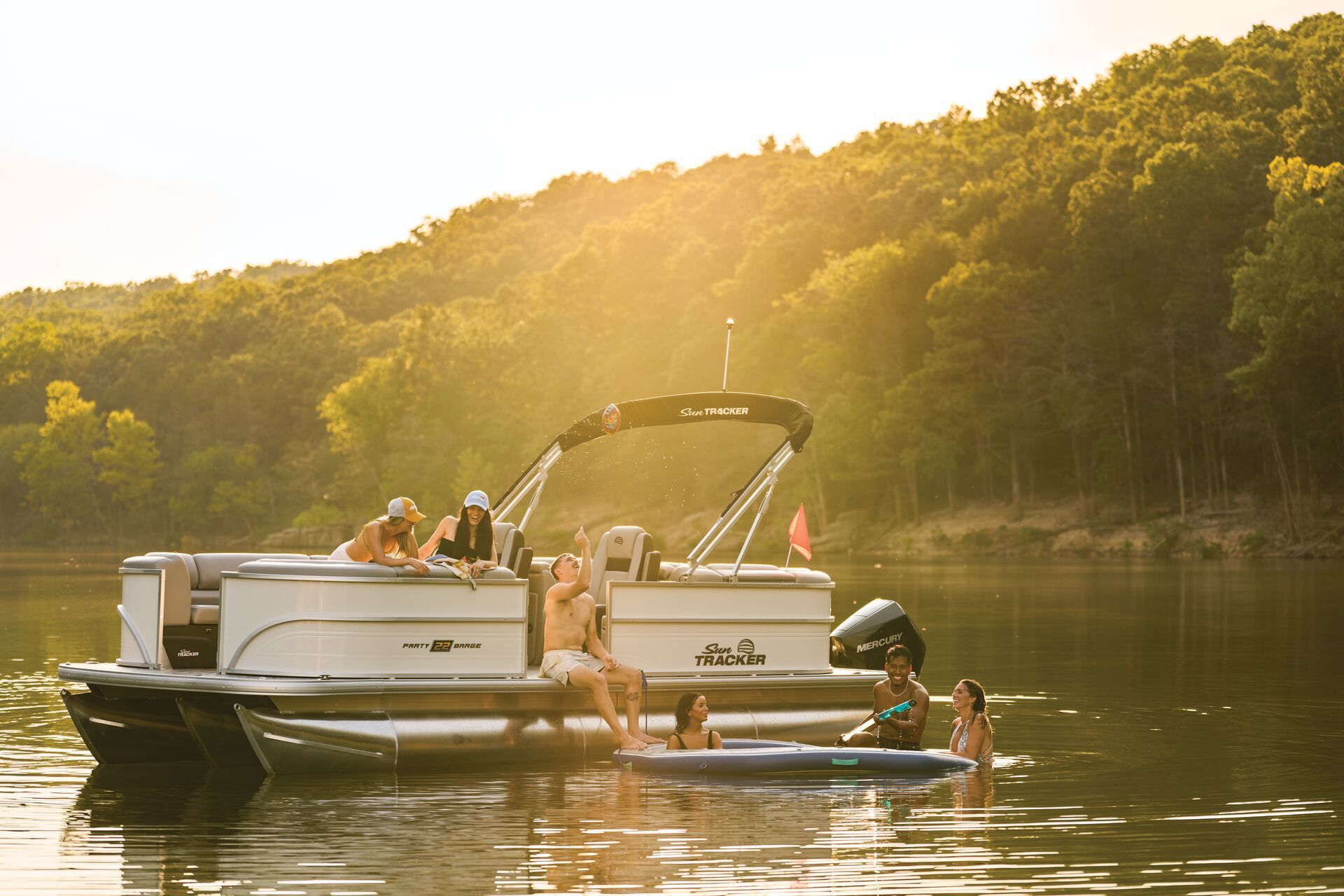 A pontoon boat filled with people sits on a calm lake at sunset, with others swimming and relaxing nearby.