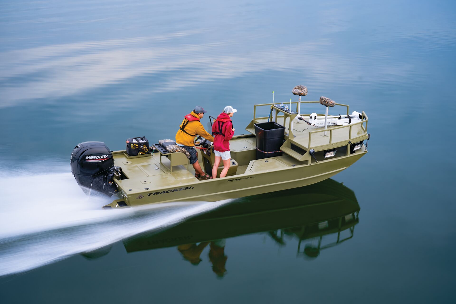 A tan utility boat moves through calm water with two people on board, creating a wake behind the motor.